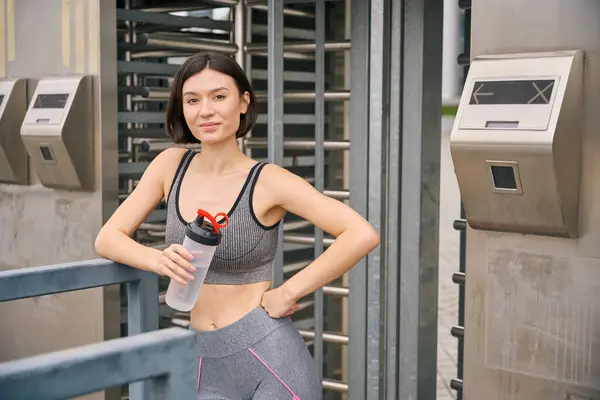 stock image Sporty young woman standing near metal constructions, holding bottle with water, resting after training. Sport concept