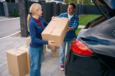 Neşeli kadın ayakta duruyor ve gülümseyen adama araba bagajında karton kutular taşıması için yardım ediyor.