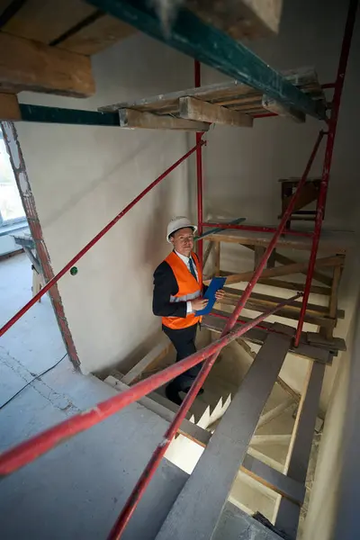 Construction inspector wearing helmet and safety vest standing on stairs while holding clipboard in hands