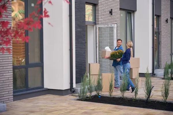 新しい家に向かって歩きながら ダンボール箱と花のポットを保持する幸せな女性を運ぶコンテンツの男 — ストック写真