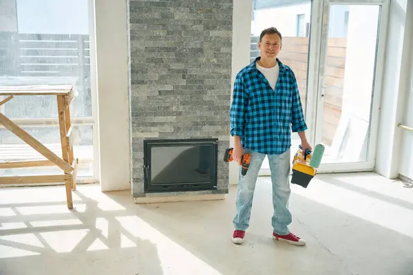 Full length photo of smiling man standing in room and holding electric screwdriver, paint roller and tool box in hands