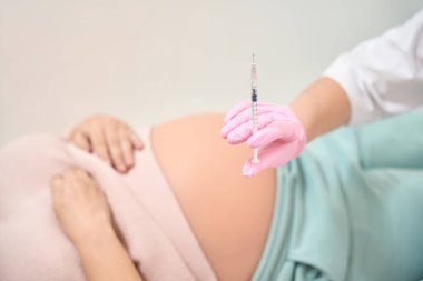 Beyaz bornoz ve güvenlik eldivenleriyle arka planda yatan sıvı, hamile bir kadın, Rh immünoglobülin enjeksiyonu, jinekoloji ve reprodükoloji.