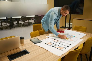 Odaklanmış genç erkek mimar çalışma alanındaki masaya yaslanırken inşaat planına kurşun kalemle bir şeyler çiziyor.