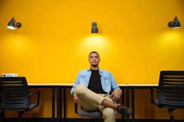 平和な若い男性のアフリカ系アメリカ人マネージャーは コールセンターの黄色い壁の近くの椅子に座って足を横切っています — ストック写真