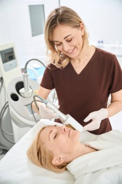 Gülümseyen doktor phlebologist, yüz derisini yumuşatmak ve damar dokusunun güçlendirilmesi ve estetik tıp kliniğinin güçlendirilmesi için genç kadının yüzüne son nokta terapisi uyguluyor.