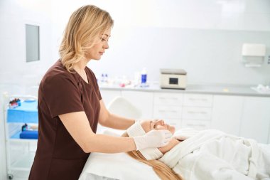 Odaklanmış kadın kozmetik uzmanı rahatlatıcı bir maske ve krem ya da serum uyguluyor cilt tipine göre kadın hasta yüzüne göre, estetik tıp kliniğinde temizlik prosedürü
