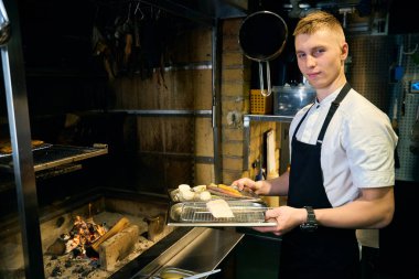 Kafkas erkek şef restoranda yanan şöminede kızarmadan önce demir tepside sebzeli pike filetosu tutarken kameraya bakıyor. Lezzetli sağlıklı yemek kavramı