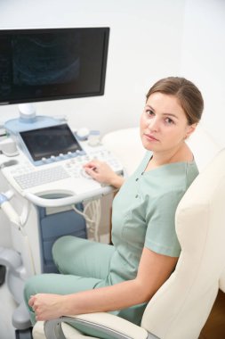 Ciddi bir beyaz kadın doktor, klinikte bulanık ultrason makinesi üzerinde çalışırken kameraya bakıyor. Sağlık hizmetleri kavramı