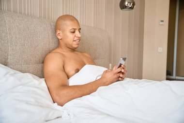 Gülümseyen ve odaklanmış bir Afrikalı Amerikalı sabah vakti otel odasında akıllı telefon kullanıyor. Dinlenme, tatil ve seyahat kavramı