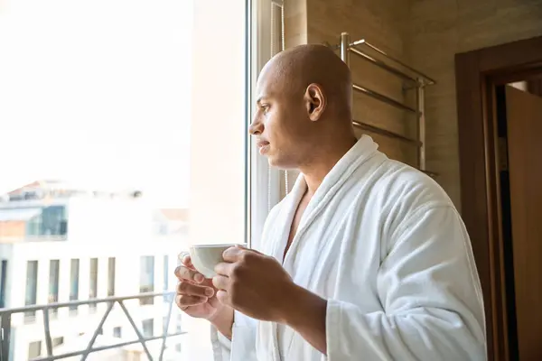 Young concentrated african american man with cup of coffee looking at window in bathroom at morning time. Concept of modern lifestyle