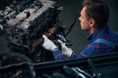 Koruyucu eldivenli genç adam araba servis merkezinde motora ekleme yapıyor.