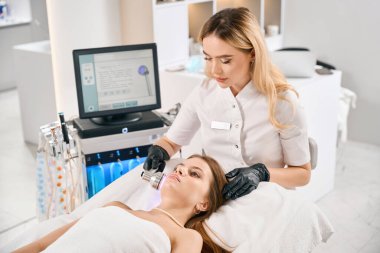 Avrupalı kadın kozmetik uzmanı bulanık güzellik salonundaki kanepede genç beyaz bir kadının yüz derisinin hafif terapisini yapıyor. Yüz bakımı, kaldırma ve gençleştirme kavramı