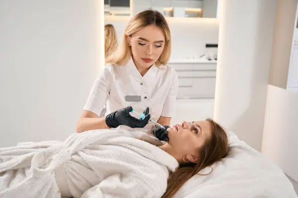 濃縮された女性のヨーロッパの化粧品学者は ぼやけた美容院でソファに若い白人女性の注射器で顔の美しさの注射を行います フェイススキンケア 持ち上がり 若返り — ストック写真