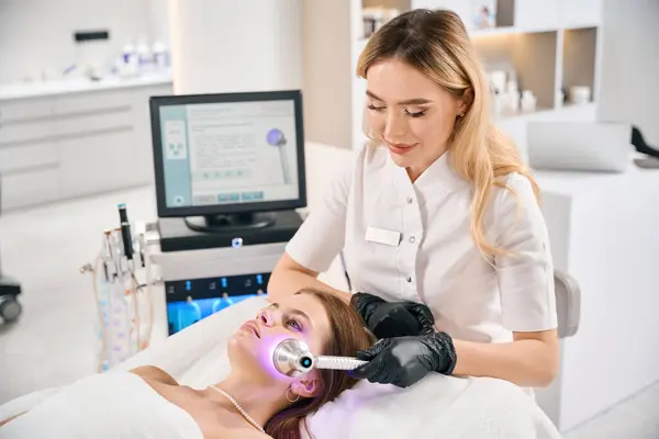 stock image Smiling female european cosmetologist doing light therapy of face skin of young caucasian woman on couch in blurred beauty salon. Concept of face skin care, lifting and rejuvenation