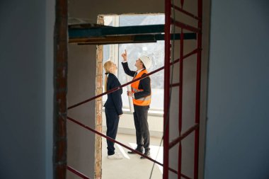 Yandan bakan erkek mimar ve kadın müşteri bitmemiş odada dikilirken yukarı bakıyor.