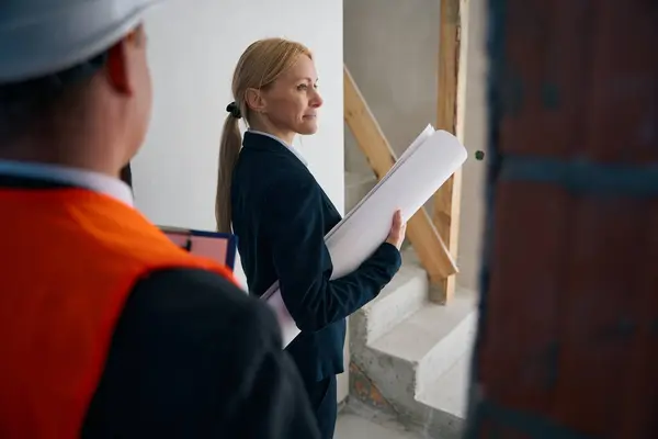 Close up photo of lady standing in house near construction foreman while holding rolled up building plan under arm