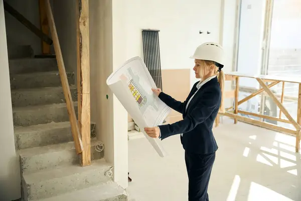 Thoughtful female construction engineer wearing helmet standing in uncompleted room and looking at building plan in her hands