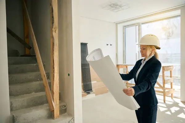 Architect lady standing in room with unfinished renewal while holding building plan in hands and looking at it