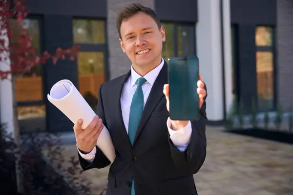 Smiling real estate broker standing near office while showing phone in hand and holding rolled up building plan under arm
