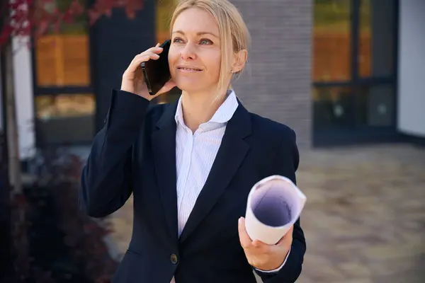 Waist up photo of cheerful female realtor standing near building while talking on phone and holding rolled up plan