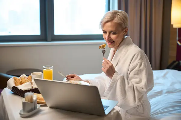 Concentrated adult caucasian woman having breakfast and watching laptop on bed at table in hotel room at morning. Concept of rest, vacation and travelling