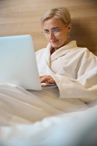 Focused adult caucasian businesswoman wearing bathrobe typing on laptop on bed in hotel room with lighting. Concept of business trip, vacation and travelling