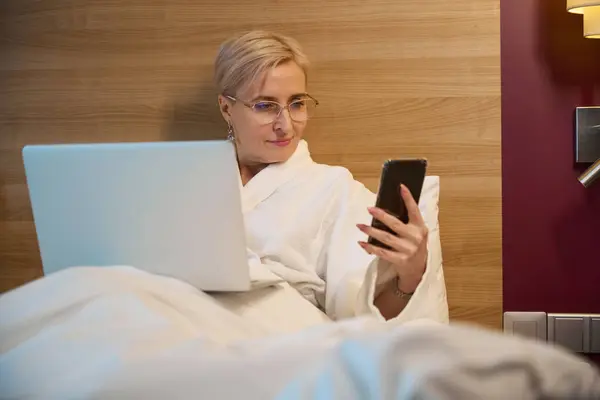 照明付きホテルの部屋のベッドで携帯電話を見ているラップトップとバスローブを身に着けている美しい大人の慎重なビジネスマン 旅行のコンセプト — ストック写真