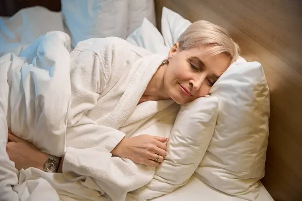笑顔の大人の白人女性は 照明付きホテルの部屋のベッドで寝るバスローブを着ています 旅行の概念 — ストック写真