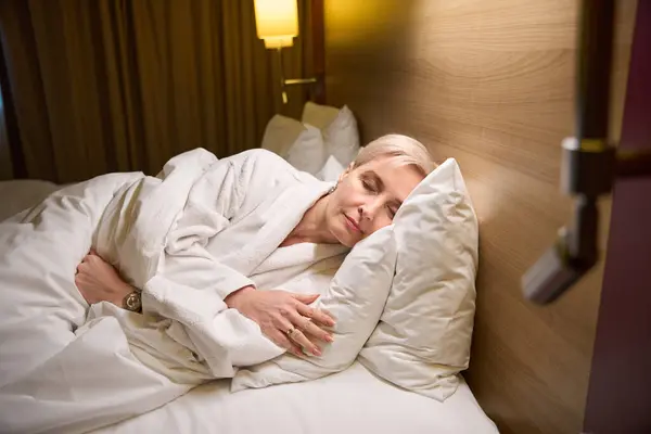 照明付きホテルの部屋のベッドで寝ているバスローブを身に着けている中年白人女性 旅行の概念 — ストック写真
