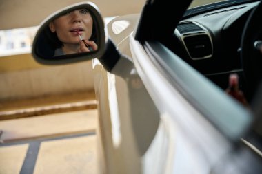 Alışveriş merkezinin otoparkında arabada otururken ruj süren kadının dikiz aynasındaki yansıması.