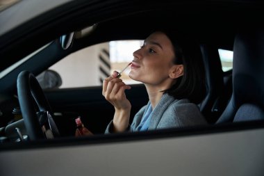Çekici bir kadın alışveriş merkezinin otoparkında otururken aynaya bakarak ruj sürüyor.