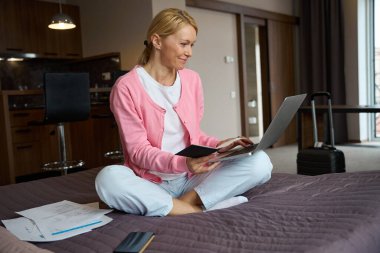 Elinde pasaport, elinde süitte oturmuş dizüstü bilgisayarla gülümseyen kadın.