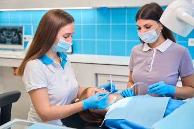 Asistanı resepsiyonda dişçiye yardım ediyor, elinde tükürük püskürtücü tutuyor, doktor genç bir hastaya diş tedavisi uyguluyor.