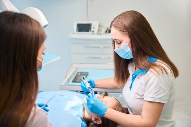 Diş asistanı elinde bir tükürük püskürtücü tutuyor, bir doktor hastasına diş matkabı takıyor.