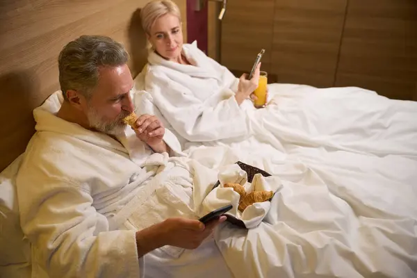 Mulher Caucasiana Adulta Olhando Para Marido Comendo Croissant Assistindo Smartphone — Fotografia de Stock