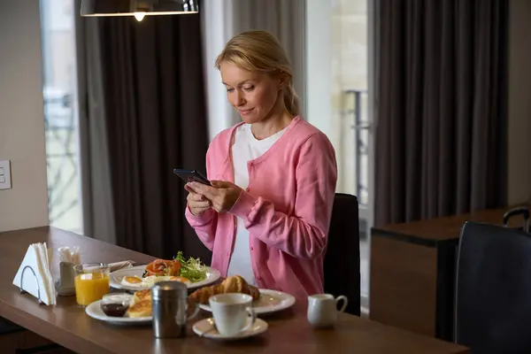 ホテルの部屋で食べ物を持ってテーブルに座っている間 彼女の手でスマートフォンを見ている集中的な女性 — ストック写真
