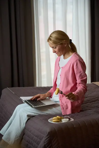 パンケーキでフォークを保持しながらコンピュータモニターを見ながらベッドに座っている笑顔の女性 — ストック写真