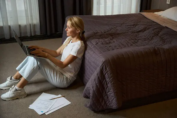 ホテルの部屋のベッドの近くのカーペットフロアに座っている間ポータブルコンピュータにタイピングする集中女性 — ストック写真