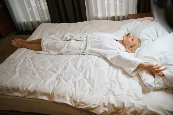 柔らかい白い枕に触れながらベッドのベッドの上でサニーに横たわるバスローブで幸せな女性 — ストック写真