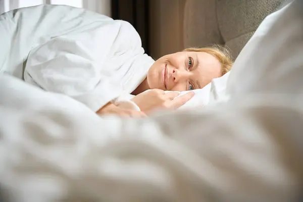 居心地の良いベッドの柔らかい枕の上に横たわる白いテリーのバスローブの笑顔の女性 — ストック写真