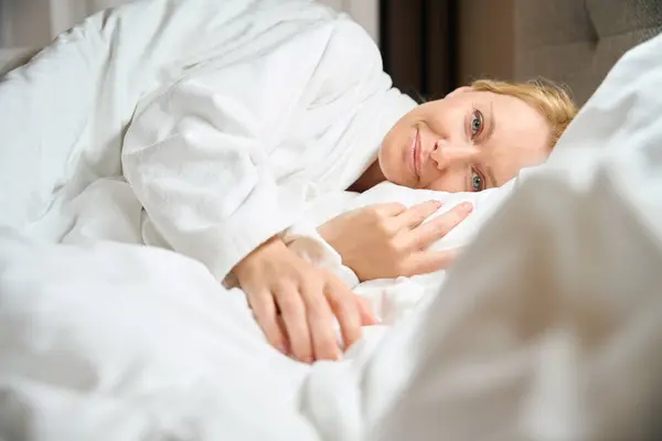 快適なベッドで柔らかい白い枕に横たわるテリーバスローブで服を着た笑顔の女性 — ストック写真
