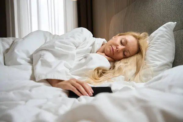 彼女の快適なベッドで数十を手で携帯電話で疲れた若い女性 — ストック写真