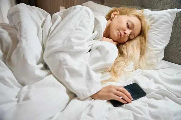 彼女の居心地の良いベッドで寝ている手のスマートフォンを持つ疲れた若い女性 — ストック写真