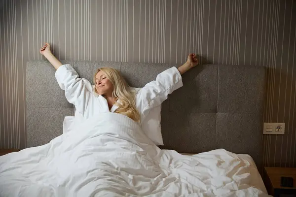 ヘッドボードに対してベッドに座っている間に伸びる白い毛布で包まれたバスローブの女性 — ストック写真