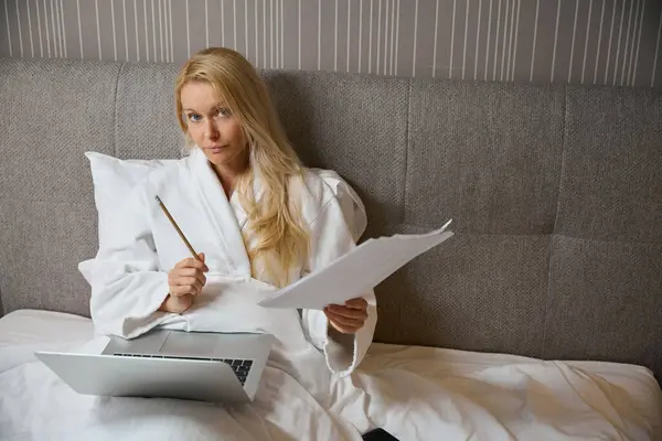 Serious businesswoman seated in bed in hotel room in front of portable computer holding pencil and documents in hands