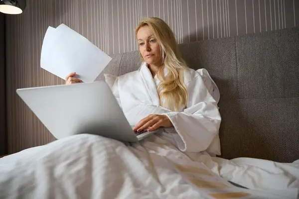 Skupiona Bizneswoman Siedząca Łóżku Apartamencie Kupą Dokumentów Ręku Pisząca Laptopie — Zdjęcie stockowe