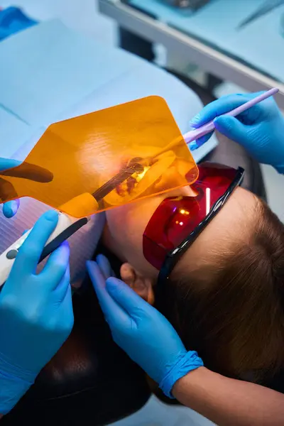 Dentist doctor uses a modern photopolymer lamp and a protective screen at work, a young female patient wears safety glasses