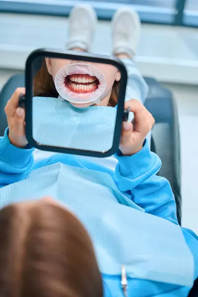 歯科オフィスの女の子は鏡で彼女の歯を見る 彼女は彼女の口に歯科トレーサーを持っています — ストック写真