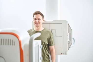 Man in upright position leaning his back against image receptor in front of x-ray machine clipart
