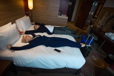 Düşünceli ve gülümseyen yetişkin beyaz iş adamı ve iş kadınının yatağında yatıp gündüz vakti otel odasına baktığı yüksek açılı manzara. İş gezisi, tatil ve seyahat kavramı